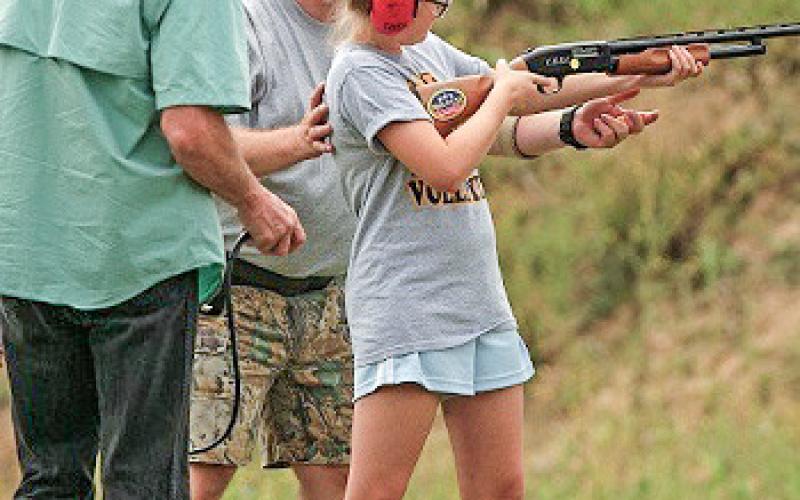 Instructors guide a student, all wearing ear protection, as she sights in a rifle outdoors.