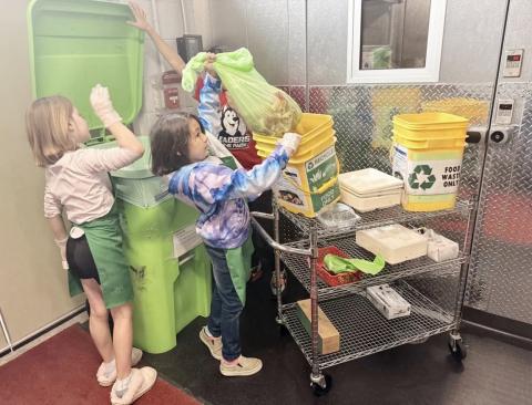 Third-graders Brooklynn Schaefer and Bella Neely place food waste into a composting bin while fourth-grader AJ Beaudion helps them by holding the lid. ISABELLA FIGUEROA NOGUEIRA/BRIDGE MICHIGAN