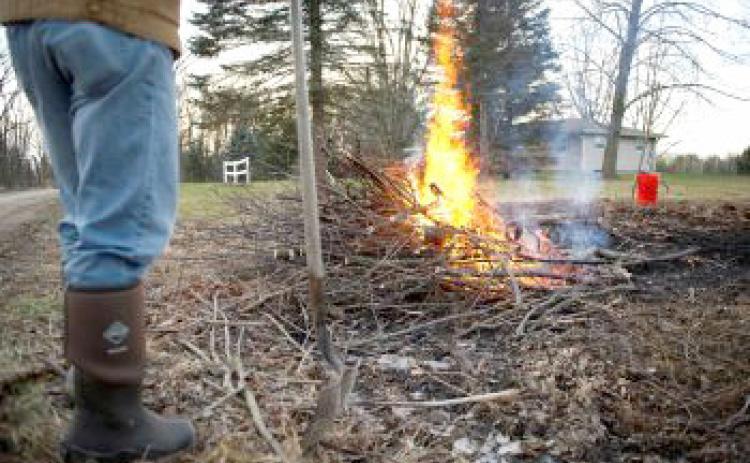 A burning brush pile is supervised by an onlooker holding a shovel.