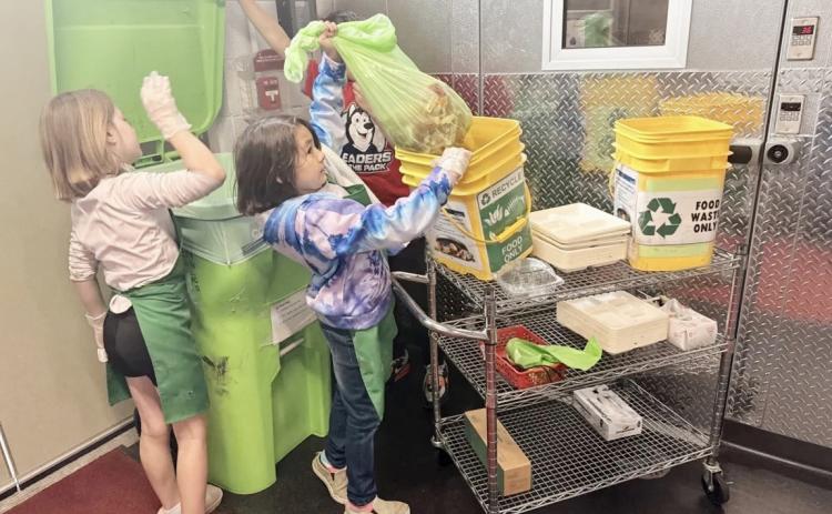 Third-graders Brooklynn Schaefer and Bella Neely place food waste into a composting bin while fourth-grader AJ Beaudion helps them by holding the lid. ISABELLA FIGUEROA NOGUEIRA/BRIDGE MICHIGAN