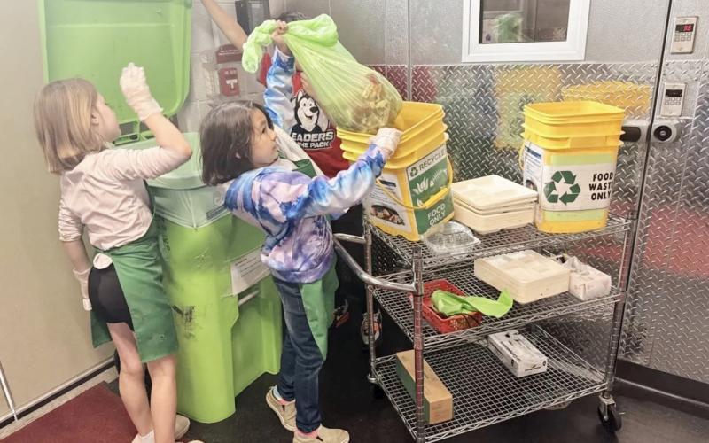 Third-graders Brooklynn Schaefer and Bella Neely place food waste into a composting bin while fourth-grader AJ Beaudion helps them by holding the lid. ISABELLA FIGUEROA NOGUEIRA/BRIDGE MICHIGAN