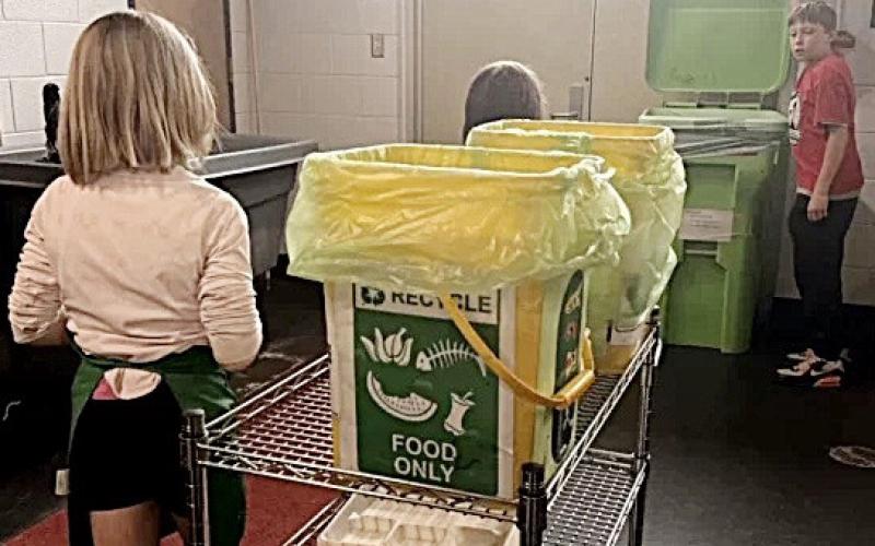 Hayes Elementary School third-graders Brooklynn Schaefer and Bella Neely roll filled food waste bins to a compost container while fourth-grader AJ Beaudion holds the lid. ISABELLA FIGUEROA NOGUEIRA/BRIDGE MICHIGAN