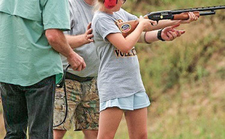 Instructors guide a student, all wearing ear protection, as she sights in a rifle outdoors.